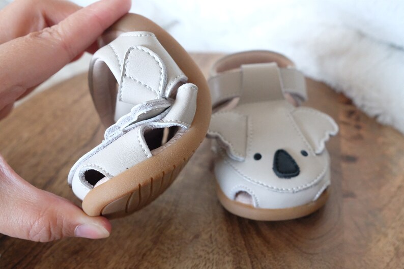 May include: Light gray baby sandals with a koala face design. The sandals have a flexible sole and adjustable straps. The koala face features black eyes and a black nose. The sandals are made of a soft material and are designed for comfort.