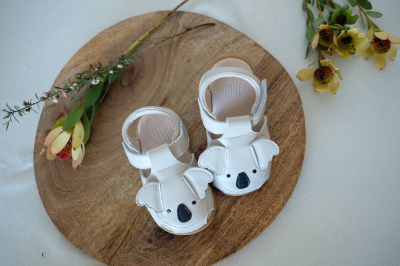 May include: A pair of white leather baby sandals with koala bear faces. The sandals have adjustable straps and are set on a wooden surface with floral accents.