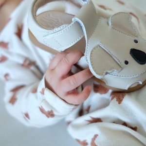 May include: A light beige baby sandal with a koala bear design on the front. The sandal has a brown sole and a velcro strap. The sandal is made of leather and is designed for infants. The sandal is approximately 4 inches long.