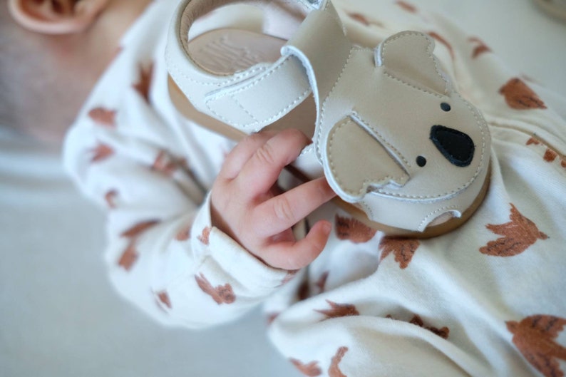 May include: A light beige baby sandal with a koala bear design on the front. The sandal has a brown sole and a velcro strap. The sandal is made of leather and is designed for infants. The sandal is approximately 4 inches long.