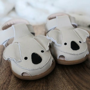 May include: A pair of light gray baby sandals with koala bear faces. The sandals have black eyes and noses, with brown soles and white stitching. The sandals are made of leather and have adjustable straps.