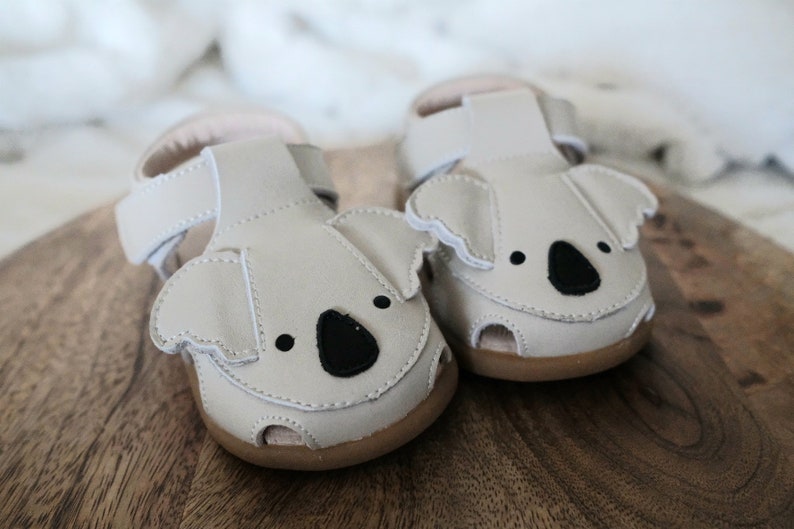 May include: A pair of light gray baby sandals with koala bear faces. The sandals have black eyes and noses, with brown soles and white stitching. The sandals are made of leather and have adjustable straps.