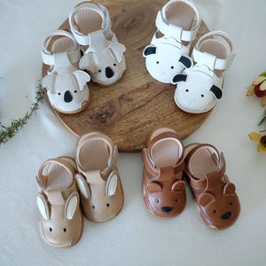 May include: A collection of baby sandals in various animal designs. The sandals include koala, sheep, bunny, and bear styles. The shoes are in neutral colors, including white, tan, and brown, with details like ears and faces.