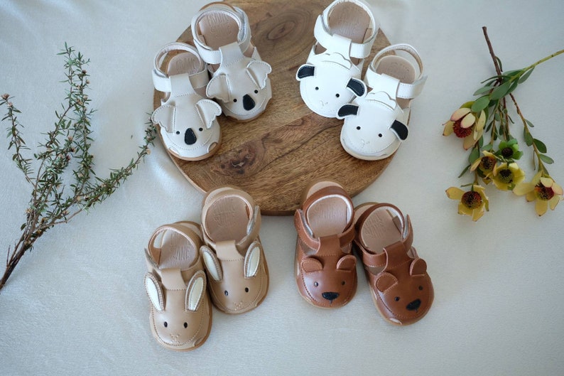May include: A collection of baby sandals in various animal designs. The sandals include koala, sheep, bunny, and bear styles. The shoes are in neutral colors, including white, tan, and brown, with details like ears and faces.