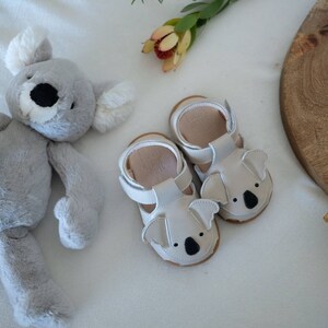 May include: A pair of baby sandals with koala bear faces, in light gray leather, sits next to a gray koala plush toy. The sandals have white straps and brown soles. A wooden serving board and flowers are in the background.