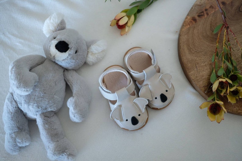 May include: A pair of baby sandals with koala bear faces, in light gray leather, sits next to a gray koala plush toy. The sandals have white straps and brown soles. A wooden serving board and flowers are in the background.