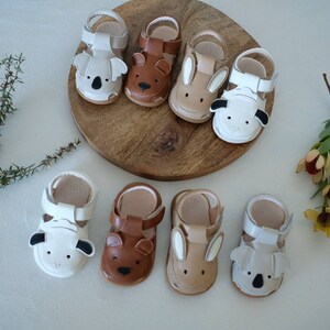 May include: A collection of baby sandals in various animal designs, including koalas, bears, and bunnies. The sandals are in neutral tones of white, brown, and tan, with detailed animal faces. They are arranged on a wooden surface.