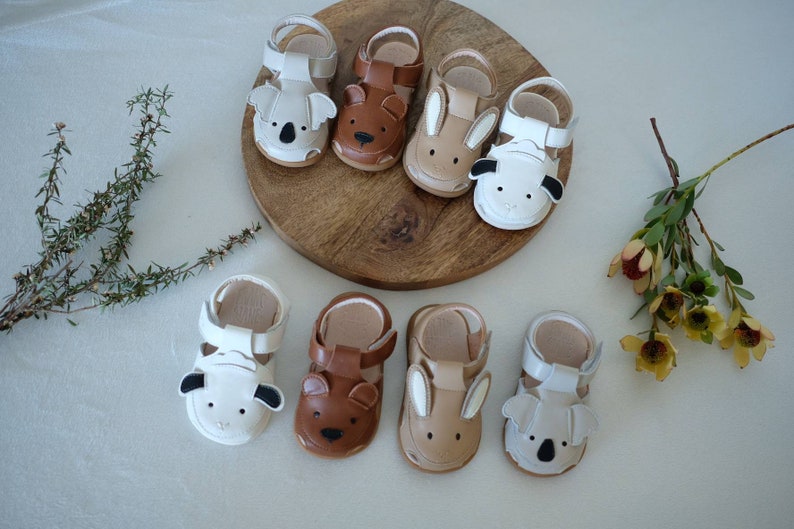 May include: A collection of baby sandals in various animal designs, including koalas, bears, and bunnies. The sandals are in neutral tones of white, brown, and tan, with detailed animal faces. They are arranged on a wooden surface.