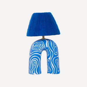 Puede incluir: Una lámpara de mesa de cerámica azul y blanca con una pantalla de lámpara de flecos azules. La base de la lámpara tiene forma de arco con un patrón de remolino.