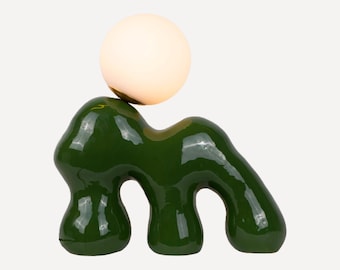 Puede incluir: Una lámpara de mesa de cerámica verde con una pantalla blanca en forma de esfera. La base de la lámpara tiene forma de animal estilizado con cuatro patas.