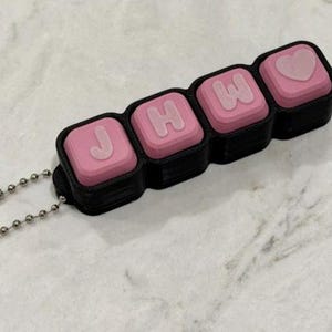May include: A black rectangular keychain featuring four square pink keys arranged in a row. The first three keys display the raised white letters J, H, and W, while the fourth key features a raised white heart symbol. A silver ball chain is attached.