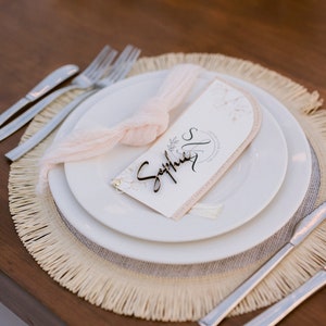 Op de afbeelding: Een witte placemat met een lichtroze stoffen servet, een houten naamschild met de naam "Sophia" erop en een witte kaart met een bloemenmotief en de tekst "Jamie & Sophia's Wedding" erop. De placemat ligt op een ronde, geweven onderzetter met franjes.