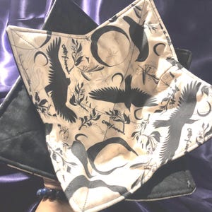 May include: A square fabric bowl cozy featuring a cream-colored interior with a black print of flying ravens, crescent moons, stars, and botanical sprigs. The exterior is solid black fabric with quilted stitching throughout the entire square structure.