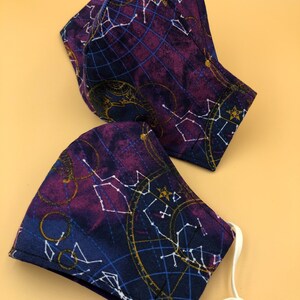 May include: Two face masks with a dark blue background and a gold and white constellation pattern. The masks have white elastic straps.