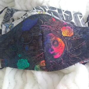 May include: Two fabric face masks with colorful floral and skull designs. The top mask is white with black and gray patterns. The bottom mask is black with colorful floral and skull designs.