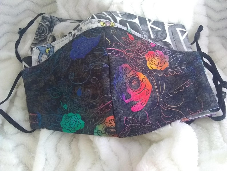 May include: Two fabric face masks with colorful floral and skull designs. The top mask is white with black and gray patterns. The bottom mask is black with colorful floral and skull designs.