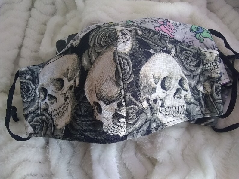 May include: Black face mask with a skull and rose design. The mask features three white skulls with black eyes and a black background with white roses.
