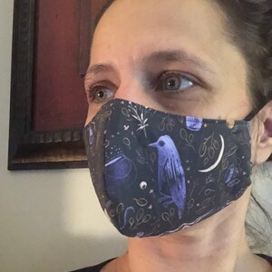 May include: A person wearing a black face mask with a purple bird, stars, and other whimsical designs.