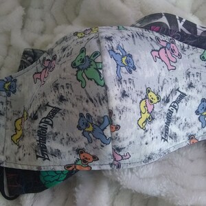 May include: A white fabric face mask with a gray and black splatter pattern. The mask features a repeating pattern of colorful dancing bears with the text "Grateful Dead" printed on the fabric.