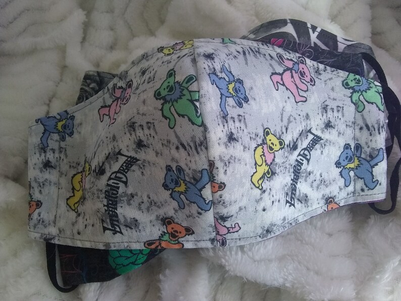 May include: A white fabric face mask with a gray and black splatter pattern. The mask features a repeating pattern of colorful dancing bears with the text "Grateful Dead" printed on the fabric.