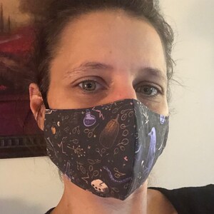May include: A person wearing a black face mask with a whimsical pattern of purple birds, eggplant, and potion bottles on a dark background.