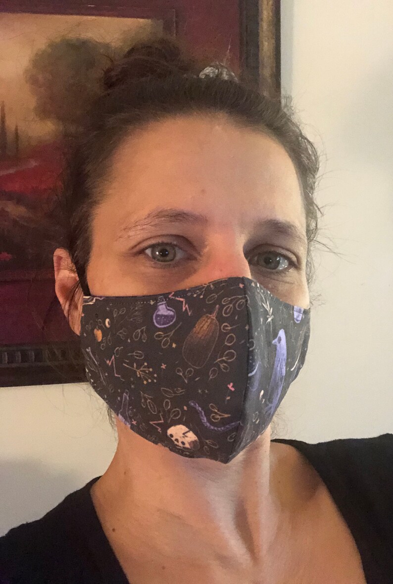May include: A person wearing a black face mask with a whimsical pattern of purple birds, eggplant, and potion bottles on a dark background.