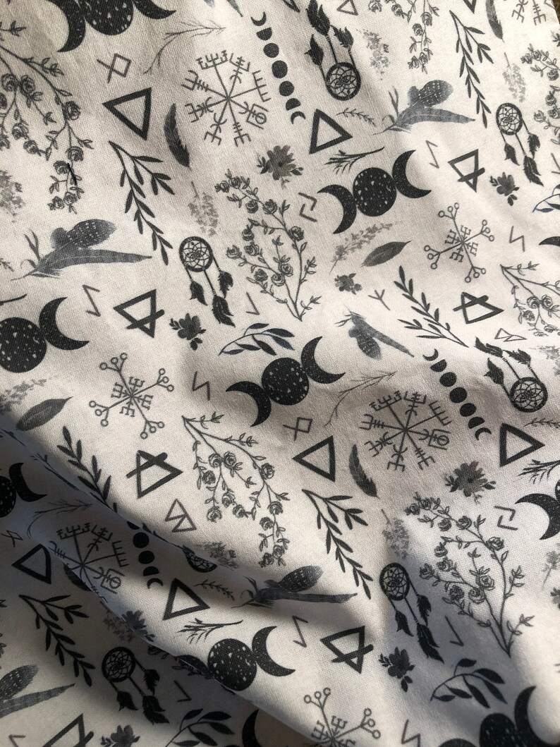 May include: A white fabric with a black and white repeating pattern of various symbols, including moons, triangles, and a compass rose. The fabric has a subtle texture.