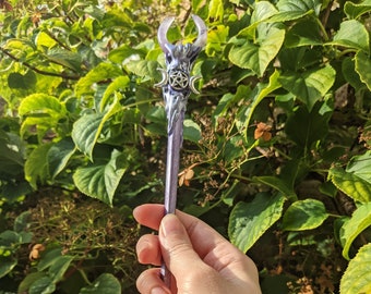 May include: A purple and silver wand with a triple moon and pentacle design at the top. The wand is made of resin and has a crystal point at the bottom.