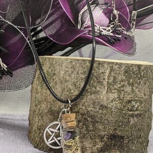 Peut inclure: Un collier en cordon de cuir noir avec un pendentif pentagramme argenté et une petite bouteille en verre remplie de cristaux, d'herbes et de sable. Le collier est suspendu à une surface en bois.