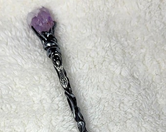 May include: A handcrafted wand with a purple amethyst crystal at the top. The wand features a black handle with silver-colored metal detailing and wire wrapping. The wand is approximately 12 inches long.