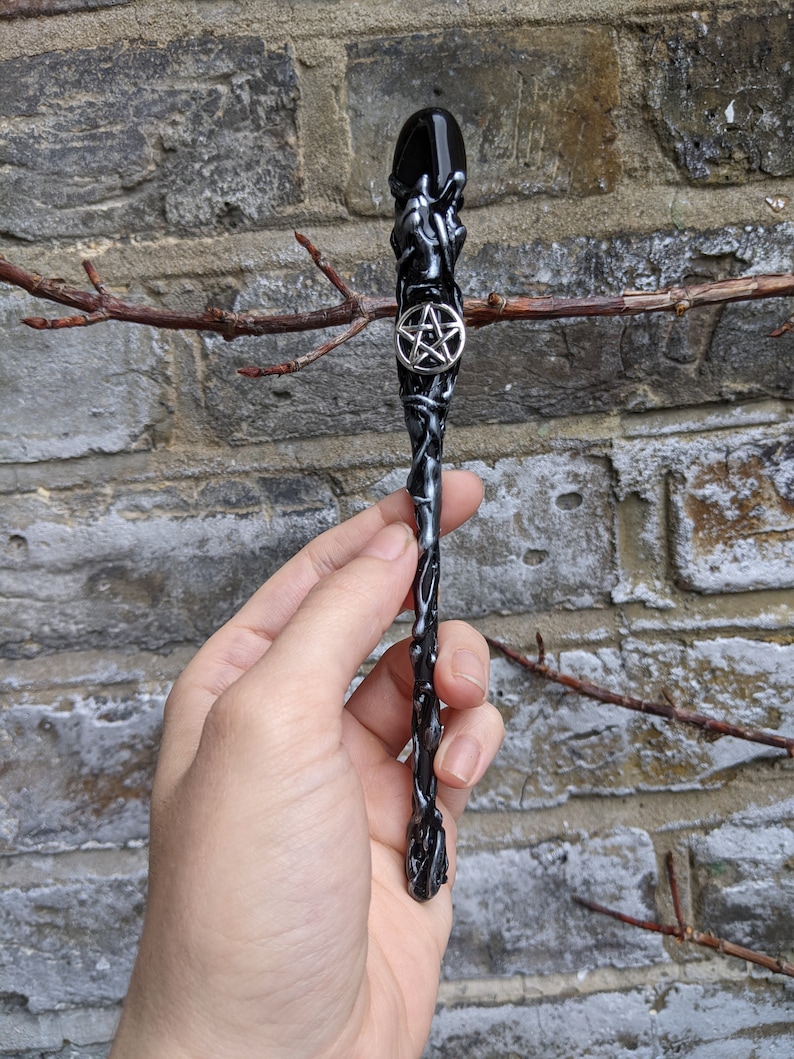 May include: Black and silver wand with a pentagram charm. The wand is made of resin and has a textured surface.