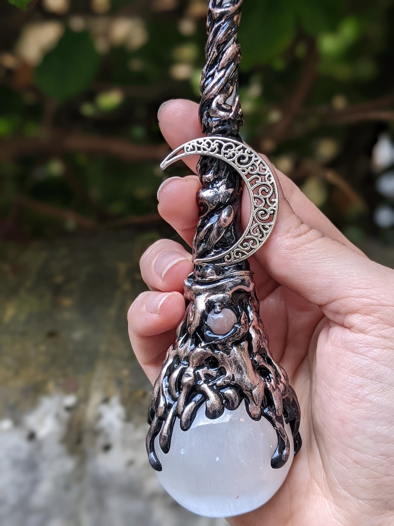 May include: A black and copper wand with a white selenite crystal orb at the end. The wand is decorated with a silver crescent moon charm.