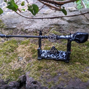 May include: A black wooden wand with a pentagram design on the handle, resting on a black wooden stand with a pentagram design. The wand is on a mossy surface.