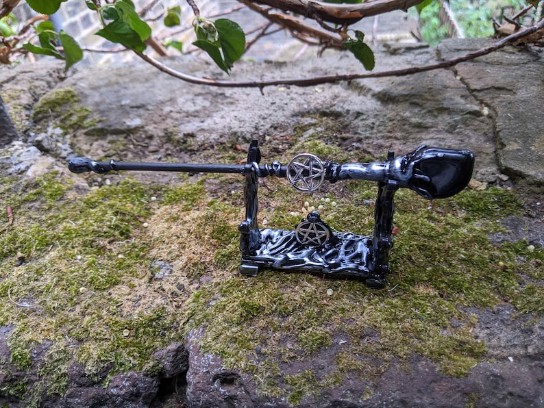 May include: A black wooden wand with a pentagram design on the handle, resting on a black wooden stand with a pentagram design. The wand is on a mossy surface.