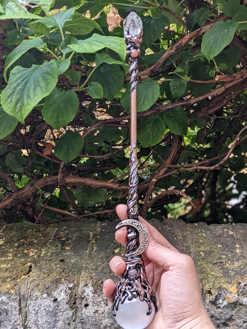 May include: A copper-toned wand with a clear quartz crystal at the top and a crescent moon charm. The wand is decorated with intricate details and has a clear quartz crystal at the base.