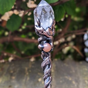 May include: A silver and copper wand with a clear quartz crystal at the top and a carnelian stone in the middle. The wand is wrapped in a vine-like design.