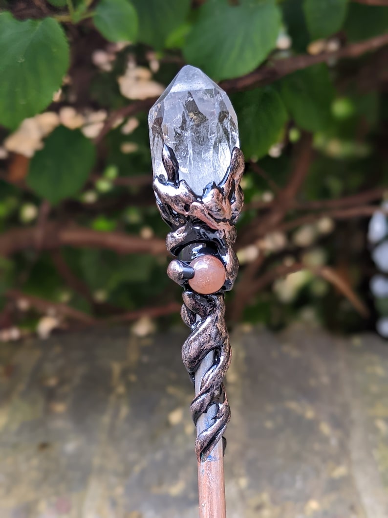 May include: A silver and copper wand with a clear quartz crystal at the top and a carnelian stone in the middle. The wand is wrapped in a vine-like design.