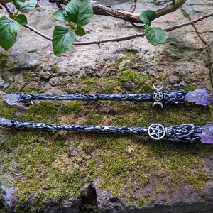 May include: Two black wands with amethyst crystal tips and silver pentagram and crescent moon charms. The wands are lying on a bed of moss.