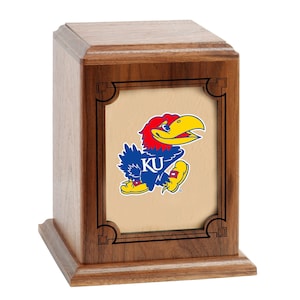 May include: A wooden cremation urn with a rectangular shape and a raised panel on the front. The panel features a brown background with a black frame and a colorful image of the University of Kansas mascot, a blue and red bird with a yellow beak and the letters "KU" on its chest.