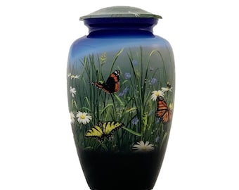 May include: A cylindrical cremation urn featuring a gradient blue to black finish. The surface displays a detailed nature scene with green grass, white daisy flowers, and three butterflies in orange, black, and yellow patterns. It has a matching lid.