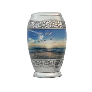 May include: A cylindrical metal cremation urn featuring a polished silver finish with intricate floral engravings on the top and bottom bands. The center displays a colorful printed scene of a sunset over the ocean with birds flying in the sky.