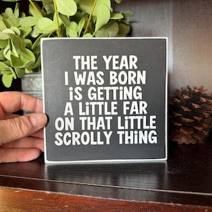 May include: A black and white square wooden sign with the text "THE YEAR I WAS BORN IS GETTING A LITTLE FAR ON THAT LITTLE SCROLLY THING". The sign is held by a hand, with greenery and pinecones in the background.