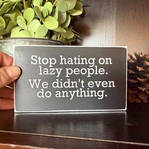 May include: A rectangular black sign with white text that reads "Stop hating on lazy people. We didn't even do anything." The sign is held in a hand, with greenery and pinecones in the background.