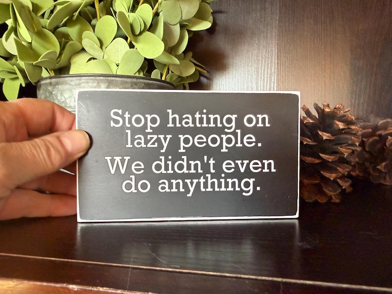 May include: A rectangular black sign with white text that reads "Stop hating on lazy people. We didn't even do anything." The sign is held in a hand, with greenery and pinecones in the background.