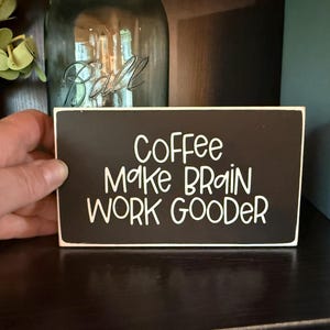 May include: A rectangular black wooden sign with white lettering that reads "Coffee Make Brain Work Gooder." The sign is held by a hand and is placed on a dark surface.