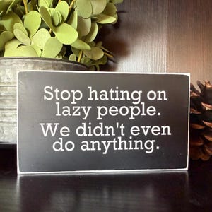 May include: A rectangular black wooden sign with white text that reads, "Stop hating on lazy people. We didn't even do anything." The sign is on a dark surface, with a metal container of greenery and a pinecone in the background.