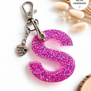 May include: A pink holographic letter S keyring featuring a silver metal lobster clasp and a small heart-shaped charm engraved with the text made with love. The letter is filled with dense pink glitter that reflects light, creating a vibrant appearance.