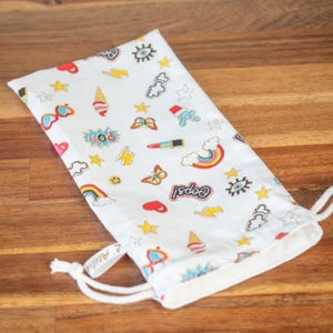 May include: White drawstring bag with a colorful pattern of rainbows, ice cream cones, stars, butterflies, and lightning bolts. The bag has the word "Oops!" printed on it and a white drawstring closure. The bag is made of fabric.