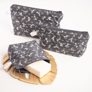 Puede incluir: Tres estuches con cremallera de color gris oscuro con un estampado de libélulas, junto a un jabonera dorada con forma de hoja que contiene una pequeña bolsa con una pastilla de jabón. El diseño de la libélula incluye libélulas blancas y detalles en rojo.
