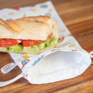 May include: A white, reusable sandwich bag with a drawstring closure, patterned with colorful rainbows, stars, and other whimsical designs. A baguette sandwich with tomato and lettuce is partially visible. The bag is on a wooden surface.